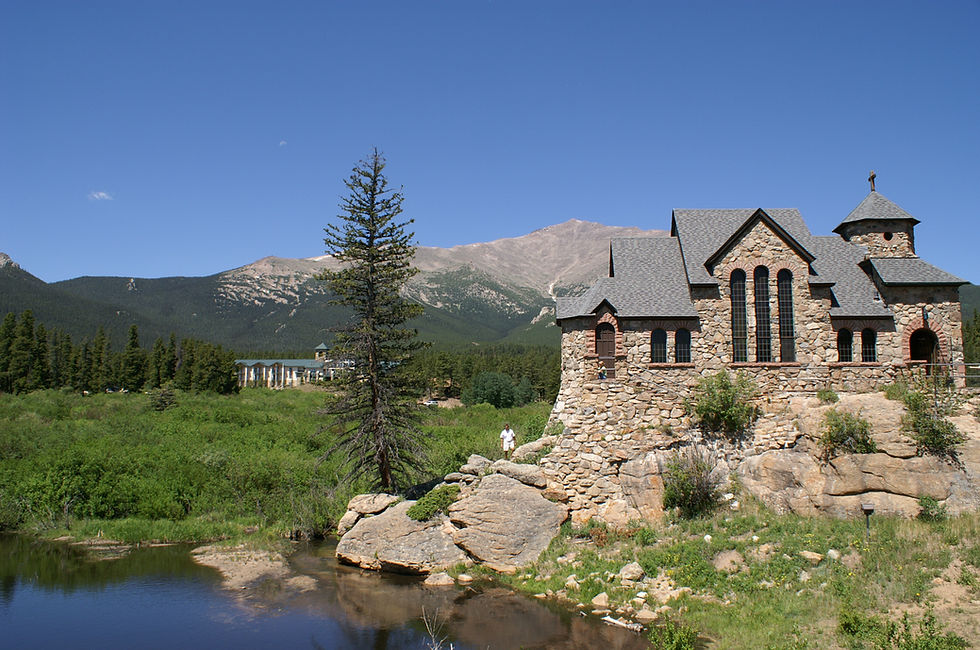 A stone chapel with steep shingled roofs and a cross stands on a rock outcrop above a grassy pond, framed by pines.